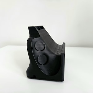 Op de afbeelding: Een matzwarte 3D-geprinte standaard met een gebogen, schuin steunvlak voor het vasthouden van een apparaat. Het zijprofiel toont een gladde, gevormde vorm met twee cirkelvormige inkepingen en een kleine rechthoekige opening aan de basis.