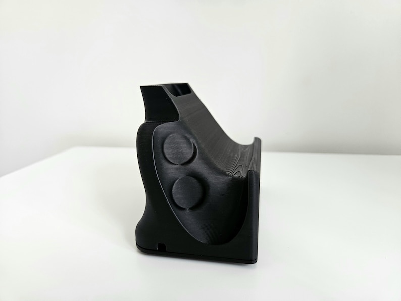 Op de afbeelding: Een matzwarte 3D-geprinte standaard met een gebogen, schuin steunvlak voor het vasthouden van een apparaat. Het zijprofiel toont een gladde, gevormde vorm met twee cirkelvormige inkepingen en een kleine rechthoekige opening aan de basis.