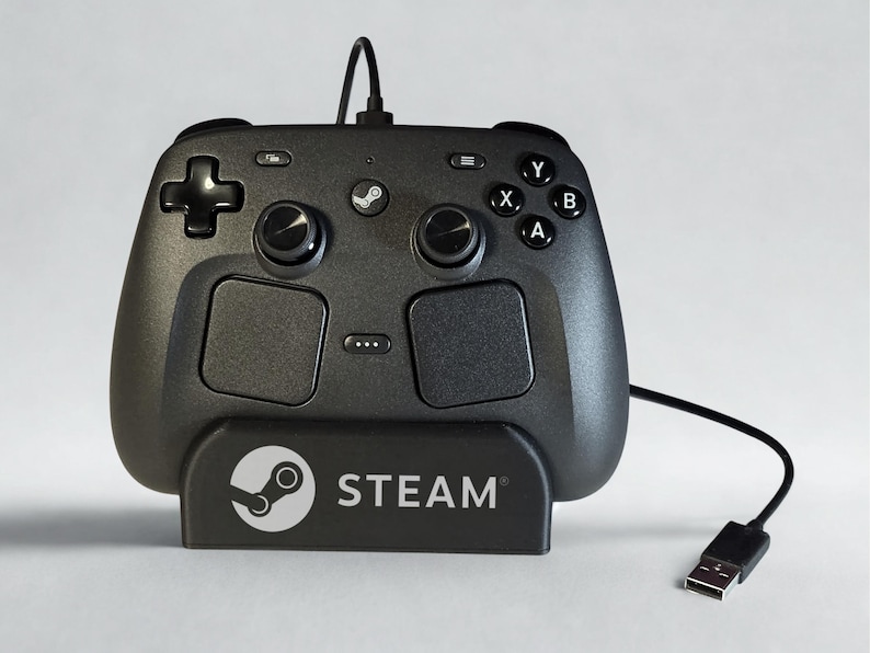 Op de afbeelding: Een zwarte Steam-controller met twee ronde trackpads, twee analoge sticks, een richtingspad en vier actieknoppen met de labels A, B, X en Y. Het apparaat is bevestigd op een zwarte standaard met het Steam-logo en de tekst Steam erop.