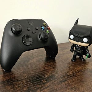 May include: A black Xbox controller with black thumbstick covers and a black Batman Pop figure on a wooden surface.