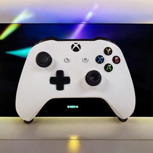 Könnte beinhalten: Weißer Xbox-Controller mit schwarzen Knöpfen und Steuerkreuz. Der Controller hat farbige Knöpfe in Grün, Blau, Rot und Gelb. Das Xbox-Logo ist oben sichtbar. Der Hintergrund ist ein dunkler Bildschirm mit bunten Streifen.