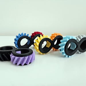 Pode incluir: Uma coleção de anéis de engrenagem em forma de infinito impressos em 3D e coloridos. Os anéis apresentam um símbolo de infinito preto com uma borda em forma de engrenagem em várias cores, incluindo azul, vermelho, amarelo, laranja, azul claro, roxo e cinza.