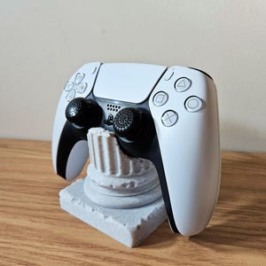 Pode incluir: Um controle de videogame branco e preto repousa sobre um suporte de pedra cinza. O controle possui joysticks e botões pretos com um direcional e botões de ação. O suporte é uma base retangular com um suporte em forma de coluna.