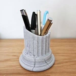 Può includere: Un portapenne grigio a forma di colonna pieno di penne di vari colori. Il portapenne è realizzato in un materiale strutturato e poggia su una superficie di legno. Le penne includono penne nere, dorate e blu.
