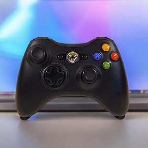 May include: Black Xbox 360 controller featuring a directional pad, two analogue sticks, and coloured action buttons. The controller has a central Xbox button, set against a blurred screen background.
