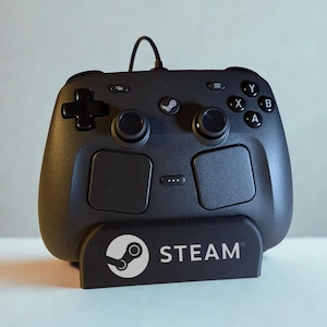 Op de afbeelding: Een zwarte Steam-controller met een richtingspad, twee analoge sticks, vier actieknoppen met de letters A, B, X en Y, en twee grote touchpads. De controller staat op een zwarte standaard met het witte Steam-logo en de tekst erop gedrukt.