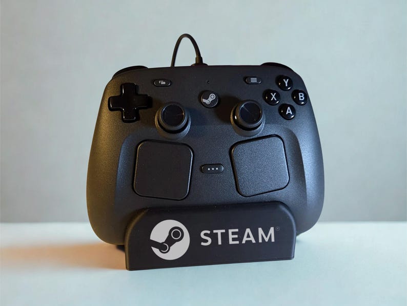 Op de afbeelding: Een zwarte Steam-controller met een richtingspad, twee analoge sticks, vier actieknoppen met de letters A, B, X en Y, en twee grote touchpads. De controller staat op een zwarte standaard met het witte Steam-logo en de tekst erop gedrukt.