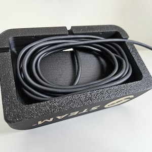 Op de afbeelding: Een zwarte rechthoekige kabelorganizer met een getextureerde afwerking en een hol middenstuk met een opgerolde zwarte kabel. De zijkant van de organizer bevat het woord STEAM in witte hoofdletters met een cirkelvormig logo-icoon.