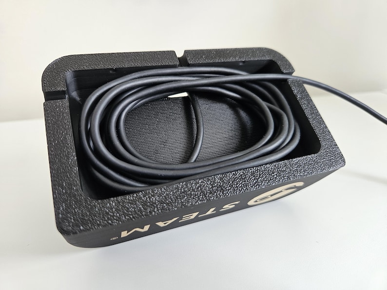 Op de afbeelding: Een zwarte rechthoekige kabelorganizer met een getextureerde afwerking en een hol middenstuk met een opgerolde zwarte kabel. De zijkant van de organizer bevat het woord STEAM in witte hoofdletters met een cirkelvormig logo-icoon.