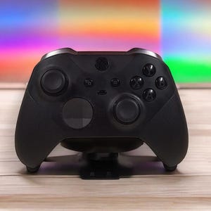 Pode incluir: Um controle Xbox preto com acabamento fosco, com dois manípulos analógicos, um direcional e vários botões. O controle está apoiado em um suporte preto, com um fundo colorido.