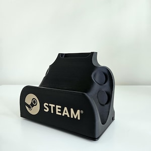 Op de afbeelding: Een zwarte plastic oplaadstandaard met het Steam-logo en de tekst in lichtbeige op de voorzijde. De standaard heeft een voorgevormde vorm met twee cirkelvormige inkepingen aan de zijkant en een uitsparing aan de bovenkant voor een apparaat.
