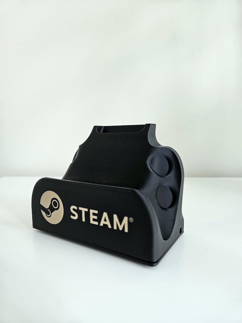 Op de afbeelding: Een zwarte plastic oplaadstandaard met het Steam-logo en de tekst in lichtbeige op de voorzijde. De standaard heeft een voorgevormde vorm met twee cirkelvormige inkepingen aan de zijkant en een uitsparing aan de bovenkant voor een apparaat.