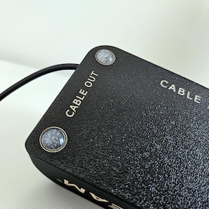 Op de afbeelding: Een zwart rechthoekig elektronisch apparaat met een getextureerd oppervlak en twee ronde grijs gespikkelde knoppen. Het bovenpaneel is duidelijk voorzien van witte tekst met de aanduidingen CABLE OUT en CABLE IN. Een dunne zwarte kabel steekt uit.