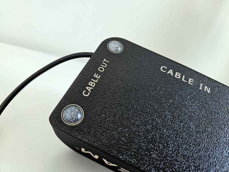 Op de afbeelding: Een zwart rechthoekig elektronisch apparaat met een getextureerd oppervlak en twee ronde grijs gespikkelde knoppen. Het bovenpaneel is duidelijk voorzien van witte tekst met de aanduidingen CABLE OUT en CABLE IN. Een dunne zwarte kabel steekt uit.
