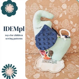 May include: A light blue and white stuffed elephant toy with a blue and white patterned fabric on the back. The elephant is sitting on a white cloud-shaped fabric with the text "IDEMpI" embroidered on it. The toy is on a brown background with a floral pattern.