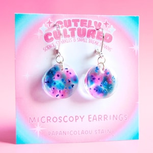 Op de afbeelding: Een paar ronde, doorzichtige hars oorbellen met een kleurrijk roze, blauw en paars gespikkeld patroon. De oorbellen zijn bevestigd aan een displaykaart met het label CUTELY CULTURED, SCIENCE, SPARKLES & SMALL BREAK DOWNS, MICROSCOPY EARRINGS, PAPANICOLAOU STAIN.