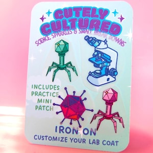 May include: A rectangular backing card featuring the text CUTELY CULTURED SCIENCE, SPARKLES & SMALL BREAK DOWNS, INCLUDES PRACTICE MINI PATCH, and IRON ON CUSTOMIZE YOUR LAB COAT. It displays four iron-on patches: a green bacteriophage, a microscope, a pink virus, and a pink bacteriophage.