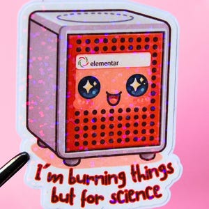 Könnte beinhalten: Ein Aufkleber mit einer cartoonartigen roten und grauen Box mit einem lächelnden Gesicht und dem Text "I'm burning things but for science." Auf der Box steht das Wort "elementar". Der Aufkleber hat ein glitzerndes Finish.