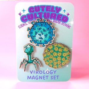 Könnte beinhalten: Ein Set aus drei Holzmagneten auf einer Karte mit der Aufschrift CUTELY CULTURED SCIENCE, SPARKLY & SMALL BREAK DOWNS und VIROLOGY MAGNET SET. Die Magnete zeigen ein blaues Virus, einen grünen Bakteriophagen und eine gelb-grüne Zelle.