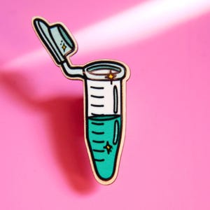 Puede incluir: Un pin con forma de tubo de ensayo de laboratorio con cuerpo blanco, marcas de medición negras y líquido turquesa. La tapa adjunta está abierta y el diseño incluye pequeños detalles de estrellas amarillas en la tapa y el líquido.