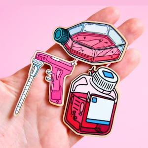 Könnte beinhalten: Drei Holz-Anstecknadeln mit Laborequipment: eine pinke Pipette, ein sechseckiger Zellkulturkolben mit pinker Flüssigkeit und eine quadratische Reagenzflasche mit weißem Etikett und pinker Flüssigkeit, alle mit schwarzen Umrandungen und Cartoon-Details.