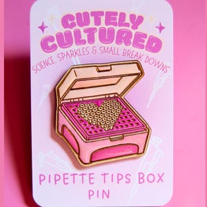 Puede incluir: Un pin de esmalte rosa que representa una caja de puntas de pipeta con una disposición en forma de corazón de puntas de pipeta. El pin tiene detalles dorados y el texto "CUTELY CULTURED" y "PIPETTE TIPS BOX PIN".