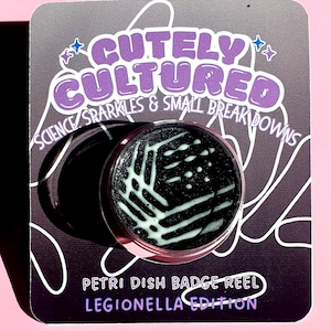 Pode incluir: Um crachá com um design de placa de Petri, com o texto "Cutely Cultured" e "Science Sparkles & Small Breakdowns". O crachá tem um fundo preto com um padrão branco e verde claro. O texto "Petri Dish Badge Reel Legionella Edition" também é visível.