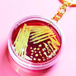 May include: A circular keychain featuring a clear plastic petri dish filled with a deep red translucent substance. Inside the dish are yellow streaks and small yellow dots resembling bacterial colonies, attached to a gold-toned metal chain.
