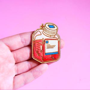 Könnte beinhalten: Holz-Anstecknadel in Form eines roten Flüssigkeitsbehälters mit weißem Etikett und Schraubverschluss. Die Anstecknadel hat eine goldene Kontur und ist vor einem rosa Hintergrund platziert. Der Behälter ist mit roter Flüssigkeit gefüllt.