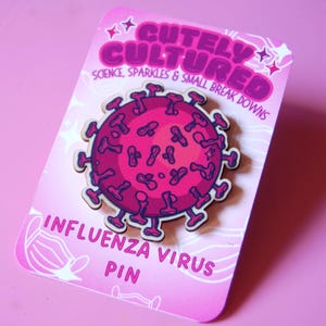 Könnte beinhalten: Eine kreisförmige Anstecknadel mit einer stilisierten magenta- und dunkelrosafarbenen Illustration eines Influenzavirus mit hervorstehenden Stacheln. Die Nadel ist auf einer hellrosa Trägerkarte mit dem Text CUTELY CULTURED und INFLUENZA VIRUS PIN befestigt.
