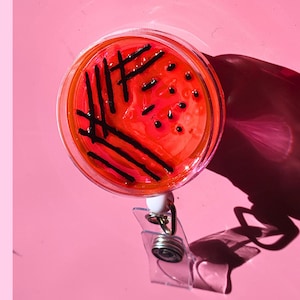 Pode incluir: Um porta-crachá redondo e transparente com um design vermelho e preto que se assemelha a uma placa de Petri. O design apresenta linhas e pontos pretos sobre um fundo vermelho. O porta-crachá tem um clipe de plástico transparente e um cordão retrátil de metal prateado.