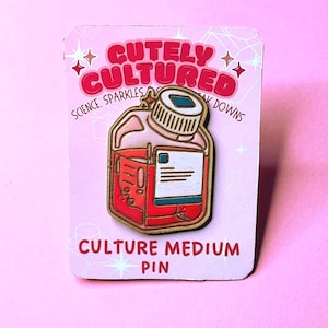 May include: A pin featuring a red culture medium flask with a white and blue label, set against a pink background. The pin is gold-colored with the words "Cutely Cultured" and "Culture Medium Pin" in red text.