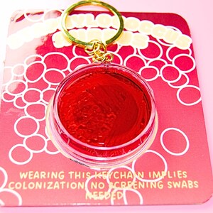May include: A circular keychain featuring a clear plastic case filled with a vibrant red gel, attached to a gold-toned metal ring. The keychain is mounted on a red backing card with white circular patterns and text that reads: WEARING THIS KEYCHAIN IMPLIES COLONIZATION NO SCREENING SWABS NEEDED.