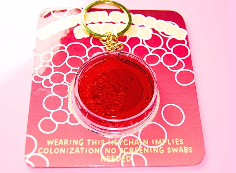 May include: A circular keychain featuring a clear plastic case filled with a vibrant red gel, attached to a gold-toned metal ring. The keychain is mounted on a red backing card with white circular patterns and text that reads: WEARING THIS KEYCHAIN IMPLIES COLONIZATION NO SCREENING SWABS NEEDED.
