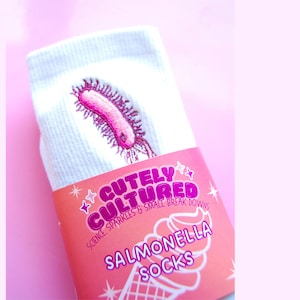 May include: White socks featuring an embroidered pink salmonella bacteria design. The packaging is a pink label with the text CUTELY CULTURED, SCIENCE, SPARKLES & SMALL BREAK DOWNS, and SALMONELLA SOCKS, along with a graphic of an ice cream cone.