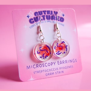 May include: A pair of round clear resin earrings featuring a red and purple Streptococcus pyogenes Gram stain pattern. The earrings hang from silver-toned metal hooks and are displayed on a pink card labeled Cutely Cultured Microscopy Earrings.