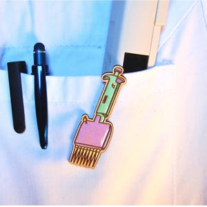 May include: A lapel pin shaped like a multichannel pipette with a light green handle and a purple base, featuring small gold star accents. The pin is attached to a white fabric pocket alongside a black stylus and a white rectangular object.