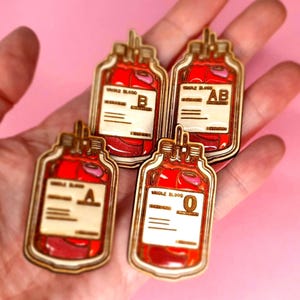 Puede incluir: Cuatro pines de madera con forma de bolsas de sangre, cada uno con una etiqueta de tipo de sangre diferente: A, B, AB y O. Las bolsas están llenas de líquido rojo y tienen un marco de madera. El fondo es rosa.