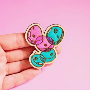 May include: A wooden pin featuring a cluster of four overlapping circular shapes. Two circles are coloured in bright pink and two are in vibrant turquoise. The design includes dark outlines and small star-shaped highlights on the surfaces.