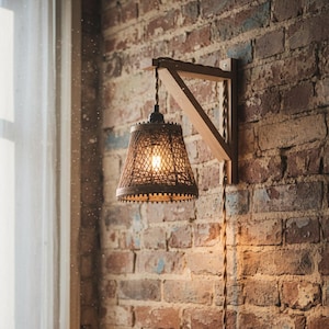 May include: A wall-mounted lamp featuring a light brown wooden bracket and a woven, conical lampshade. The lamp is suspended by a twisted fabric cord that runs down the wall, and it emits a warm, glowing light from within the shade.