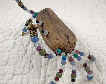 May include: A decorative sculpture featuring a central piece of brown driftwood adorned with multiple strands of colorful beads. The beads are made of various materials in shades of blue, green, purple, and clear, accented with ornate gold-tone metal caps.