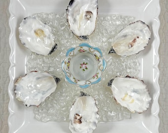 May include: A square white scalloped serving platter holds five iridescent oyster shells arranged around a central small porcelain bowl. The bowl features a light blue rim, gold trim, and a delicate floral pattern with pink roses and green leaves.