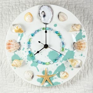 May include: A round white clock with a coastal theme. The clock face is decorated with various seashells, a starfish, and a ring of blue and green beads. Teal paint splatters add a decorative touch. The clock hands are black.