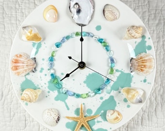May include: A round wall clock with a white face featuring teal paint splatters. The hour markers are represented by various natural seashells and a starfish at the bottom. A circle of blue and green glass pebbles surrounds the black clock hands.