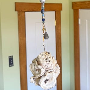 May include: A hanging ornament featuring a natural oyster shell. The shell is cream-colored with a textured surface. It is suspended by a wire and adorned with a string of blue, gold, and gray beads. The ornament hangs in front of a white door.