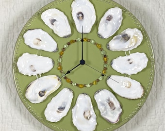 May include: A round wall clock with a sage green face featuring a beaded border. Twelve white oyster shells are arranged as hour markers around the center. A circle of small amber and light green beads surrounds the black clock hands in the middle.