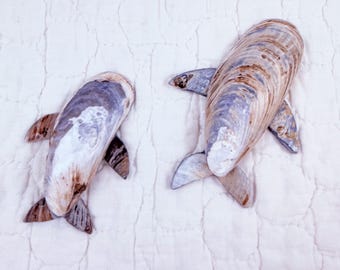 May include: Two decorative figures shaped like dolphins, crafted from natural seashells. The shells feature a smooth, polished surface with swirling patterns in shades of brown, tan, gray, and white, mimicking the sleek form of aquatic mammals.