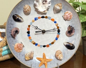 May include: A decorative clock with a white face and a blue-gray rim. The clock features seashells and colorful stones around the face, with a starfish at the bottom. The clock hands are black.