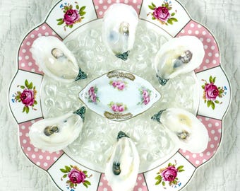 May include: A round ceramic plate with a scalloped edge featuring pink polka dots and pink rose floral patterns. The plate holds a bed of clear ice cubes, five raw oysters, and a small, oval-shaped white ceramic dish with pink rose accents.
