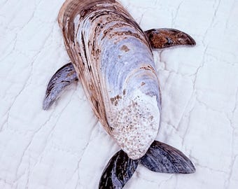 May include: A decorative sculpture shaped like a whale, constructed from various natural seashells. The body is a large, elongated brown and tan shell, while the fins and tail are composed of smaller, darker, polished grey and black shells.